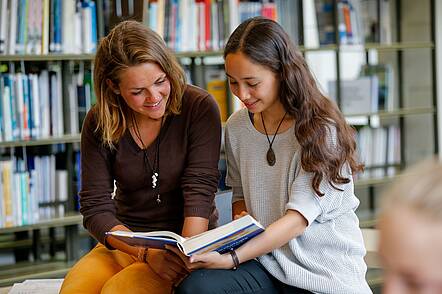 Foto: Zwei Studentinnen sitzen in der Bibliothek und lesen in einem Buch.